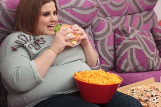 A fat women lounging on a sofa eating a hamburger and other snacks